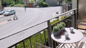 A balcony with a table and chairs, overlooking a road with a pedestrian crossing and parked cars. - Cozy apartment facing south - 4 guests (Saint-Lary-Soulan)