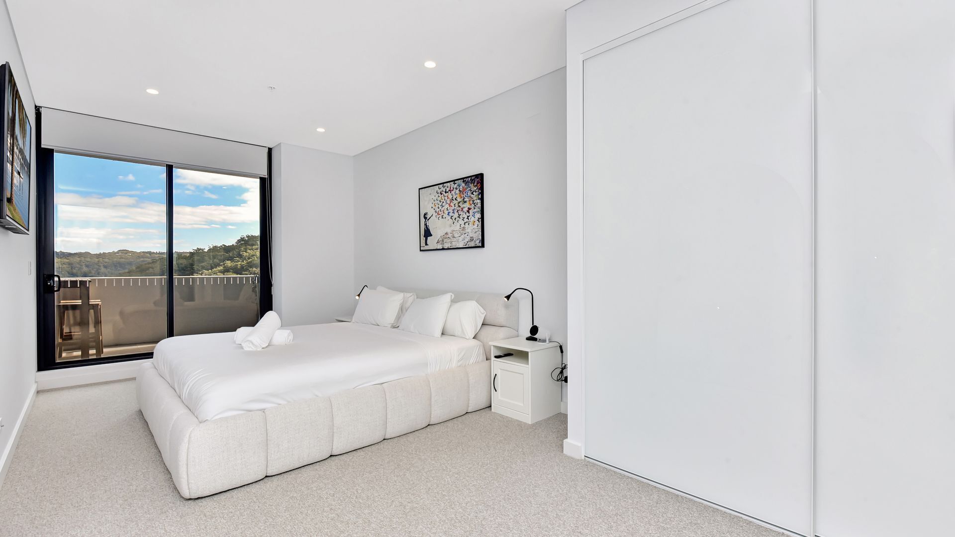 A modern bedroom with a large bed, a bedside table, a nightstand, and a window with a view of the outdoors.