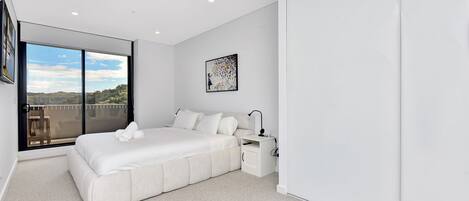 A modern bedroom with a large bed, a bedside table, a nightstand, and a window with a view of the outdoors.