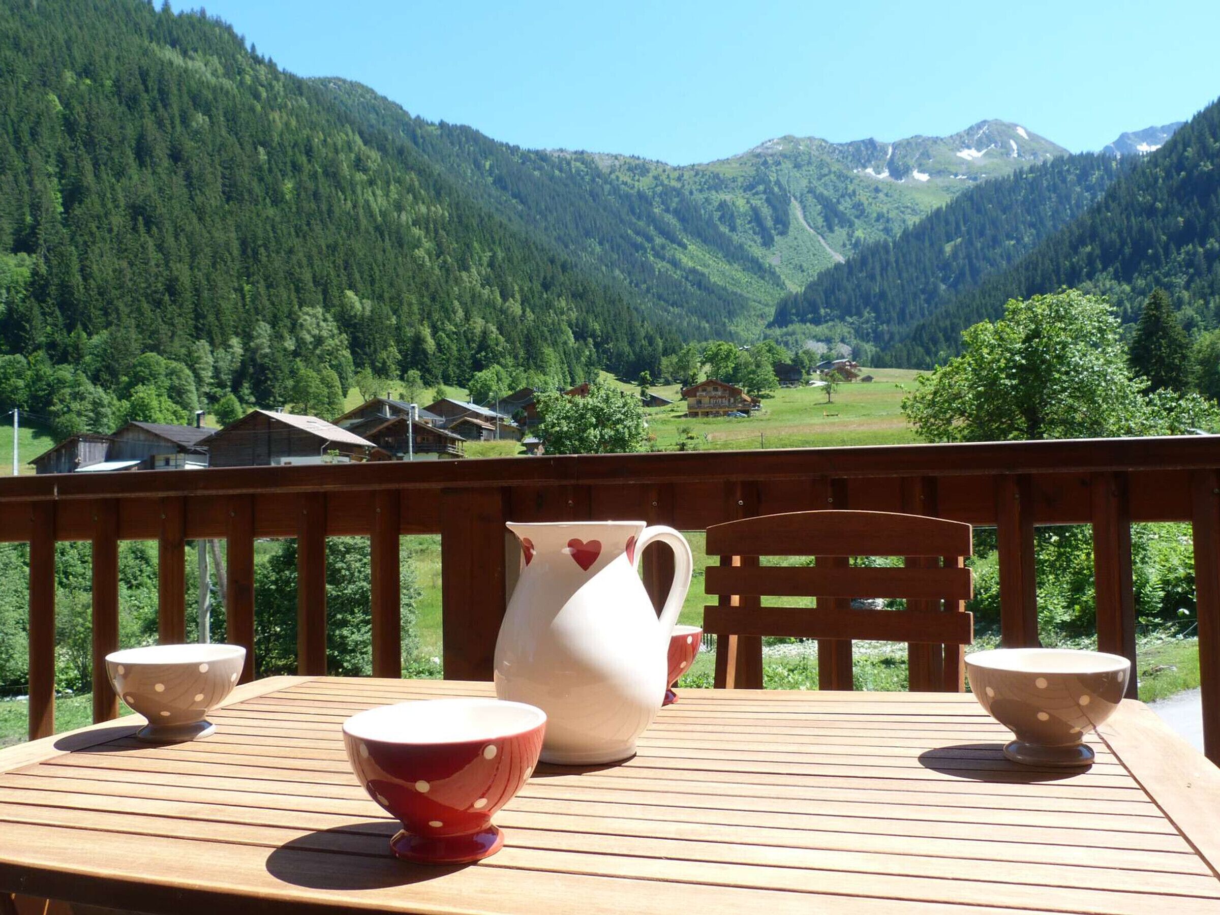 Eine Holzterrasse mit Tisch, zwei Schüsseln und einer Karaffe, mit Blick auf eine gebirgige Landschaft mit Häusern.