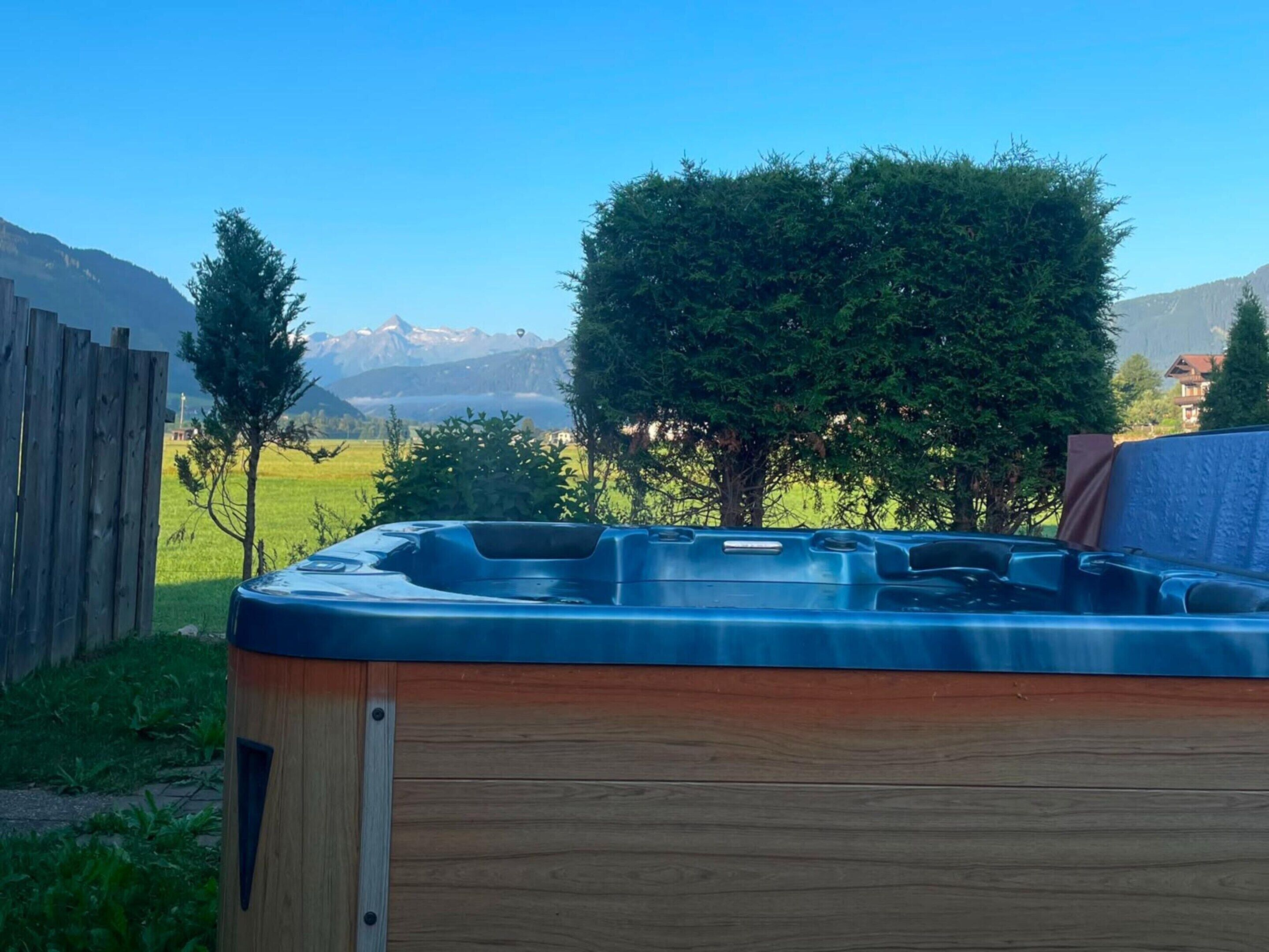 Un jacuzzi con base de madera y tapa metálica, rodeado de un cerco de madera y árboles, con una vista de la montaña al fondo.
