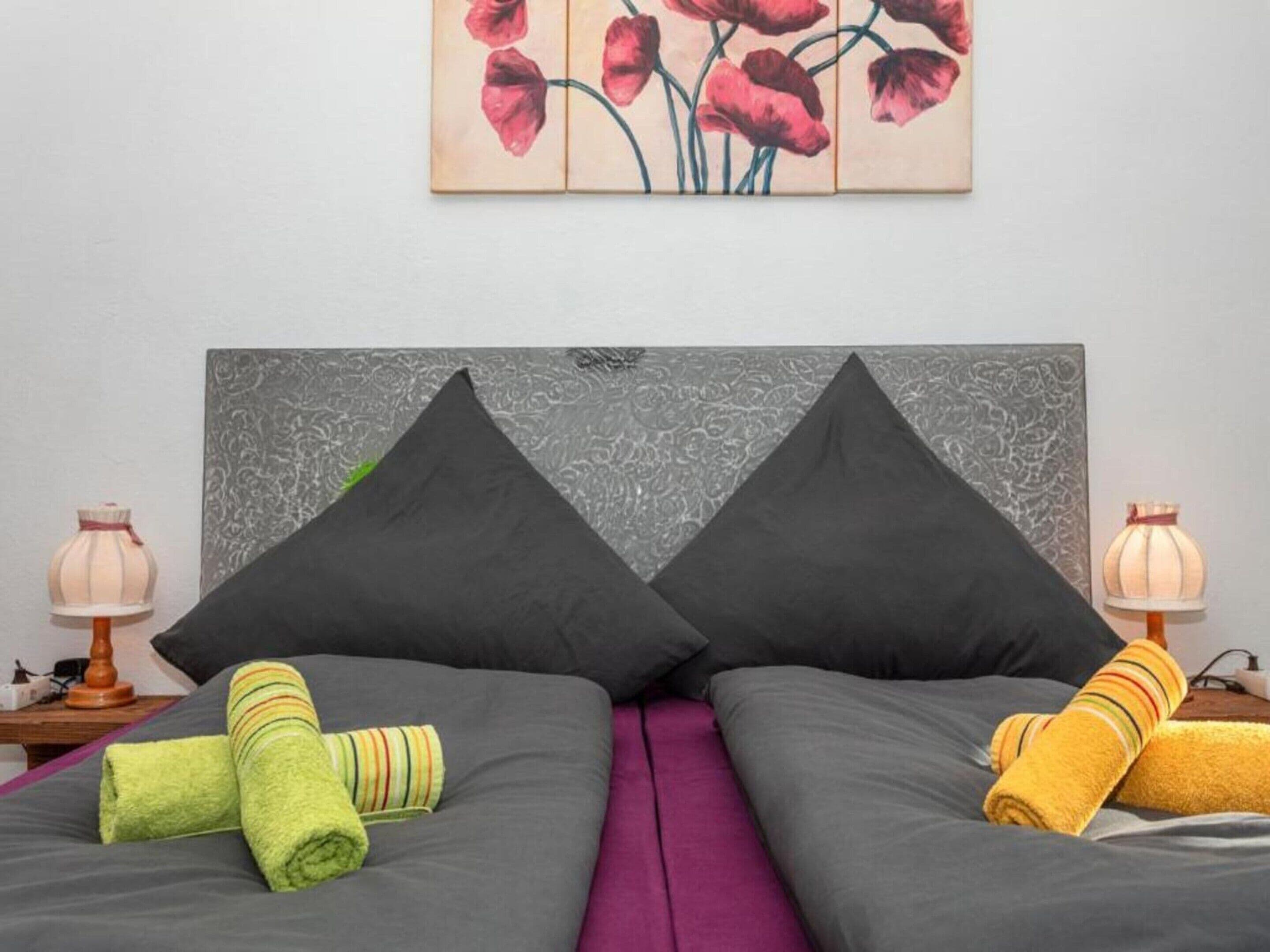 Una cama con dos almohadas, una toalla verde y otra amarilla, y un cuadro de flores rojas en la pared.