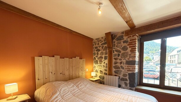 A bedroom with a bed, a nightstand, a lamp, a stone wall, and a window with a view of parked cars and buildings. - Charming house, parking, city center - 8 guests (Mont-Dore)