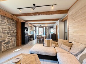 A modern living room with a sectional sofa, a dining area, and a kitchen with stone walls. - Charming house, parking, city center - 8 guests (Mont-Dore)