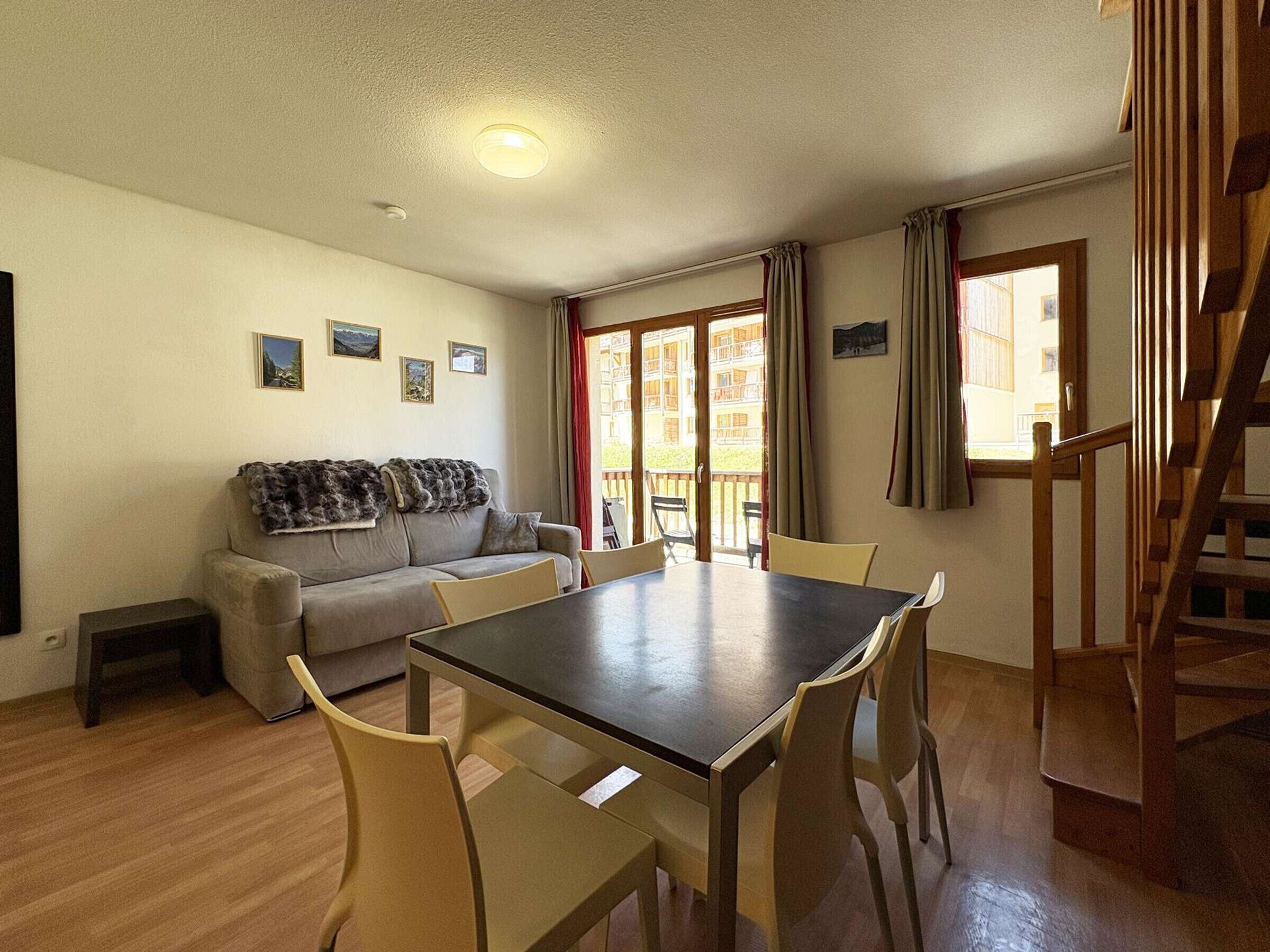 A living room with a sofa, a dining table with chairs, a staircase, and a view of the outside.