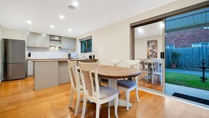 A modern kitchen with a dining area, wooden flooring, and a sliding glass door leading to a backyard. - The Burwood Cottage Comfort & Convenience (Burwood)