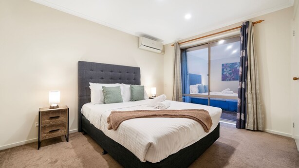 A hotel room with a large bed, a bedside table with a lamp, a wall-mounted air conditioner, and a view of the outside through a glass door. - The Burwood Cottage Comfort & Convenience (Burwood)
