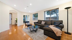 A spacious living room with a dark grey sofa, a coffee table, and a large window providing a view of the outdoors. - The Burwood Cottage Comfort & Convenience (Burwood)