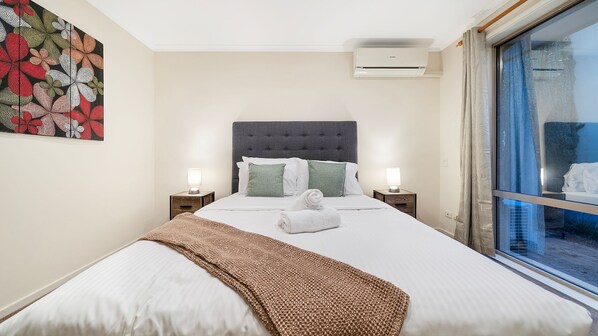 Habitación de hotel con cama, dos mesitas de noche, aire acondicionado de pared y ventana con cortinas.