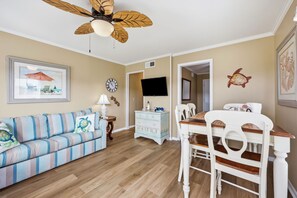 Ein Zimmer mit einem gestreiften Sofa, einem Holzesstisch mit Stühlen, einem Deckenventilator, einer Uhr und einem gerahmten Bild an der Wand.