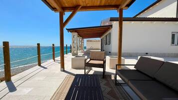 Un área de estar al aire libre techada con vistas al mar, una pérgola de madera y un edificio blanco al fondo.