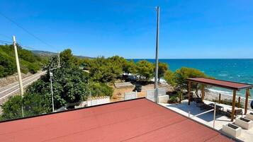 Una terrazza con tetto in tegole rosse e posti a sedere all'aperto, con vista su un paesaggio costiero con alberi e cielo azzurro limpido.