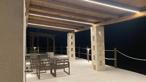 Een overdekt terras buiten met houten banken en een pilaarstructuur van natuursteen.