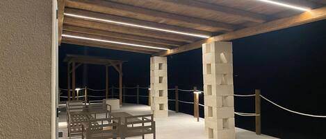 Un espace extérieur couvert, aménagé en patio, avec des bancs en bois et une structure en piliers de pierre.