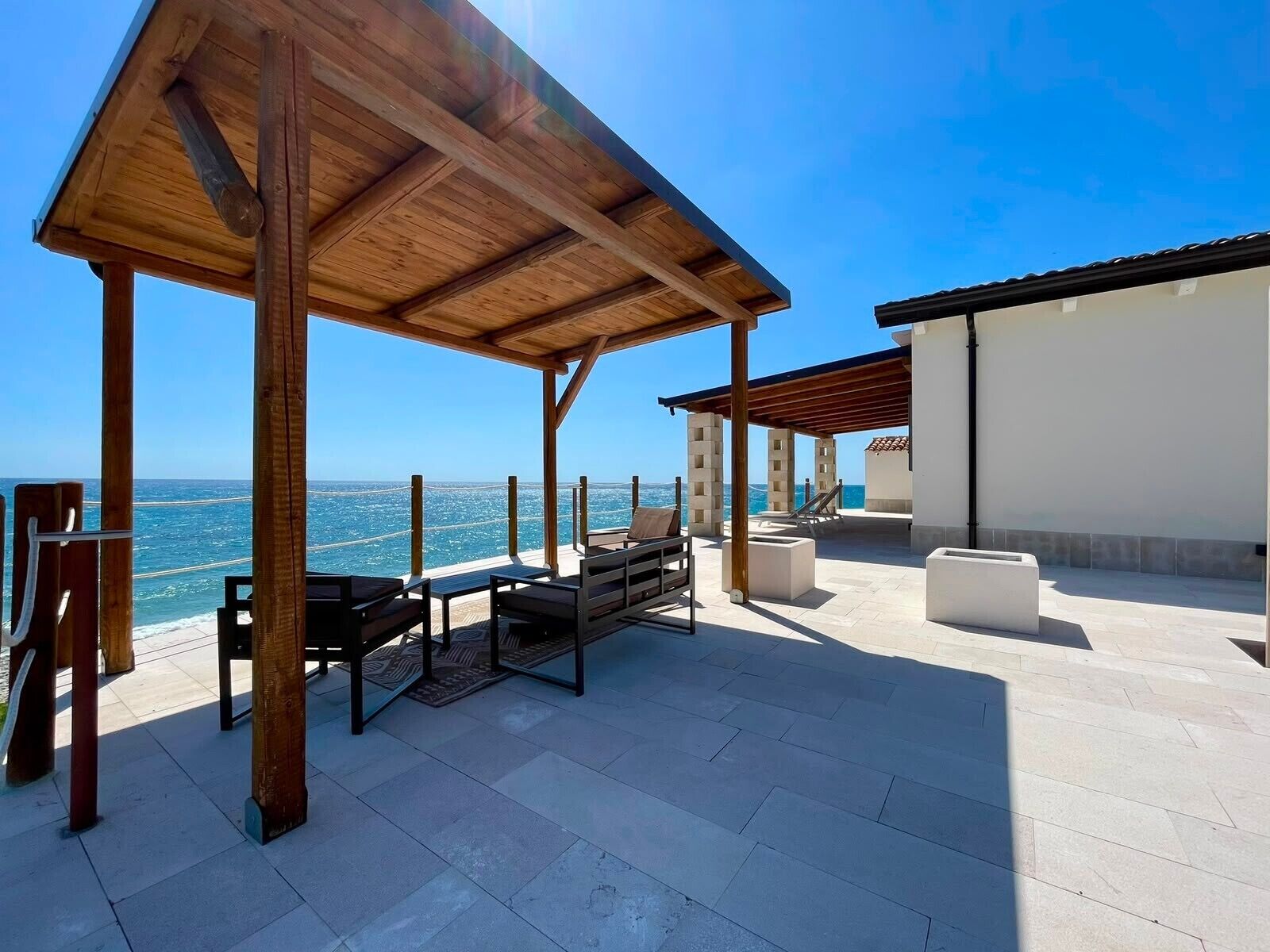 Un'area salotto esterna coperta con vista sul mare, caratterizzata da un pergolato in legno, poltrone relax e un edificio bianco sullo sfondo.