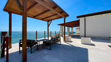 Un'area salotto esterna coperta con vista sul mare, caratterizzata da un pergolato in legno, poltrone relax e un edificio bianco sullo sfondo.