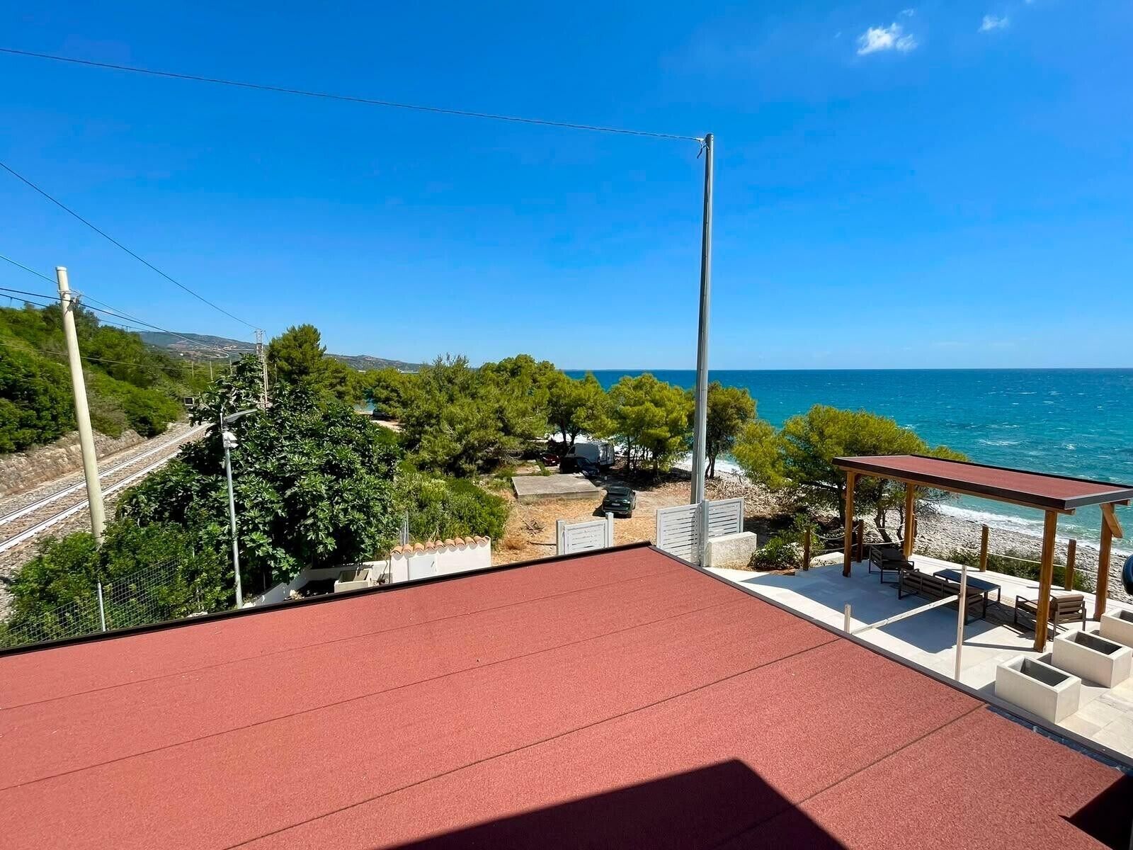 Une terrasse sur le toit, recouverte de tuiles rouges, avec des sièges d’extérieur, offrant une vue sur un paysage côtier agrémenté d’arbres et d’un ciel bleu dégagé.