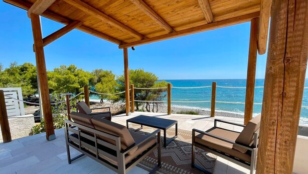 Ein Sitzbereich im Freien mit einer Holzpergola, Blick auf das Meer und einem Strand im Hintergrund.