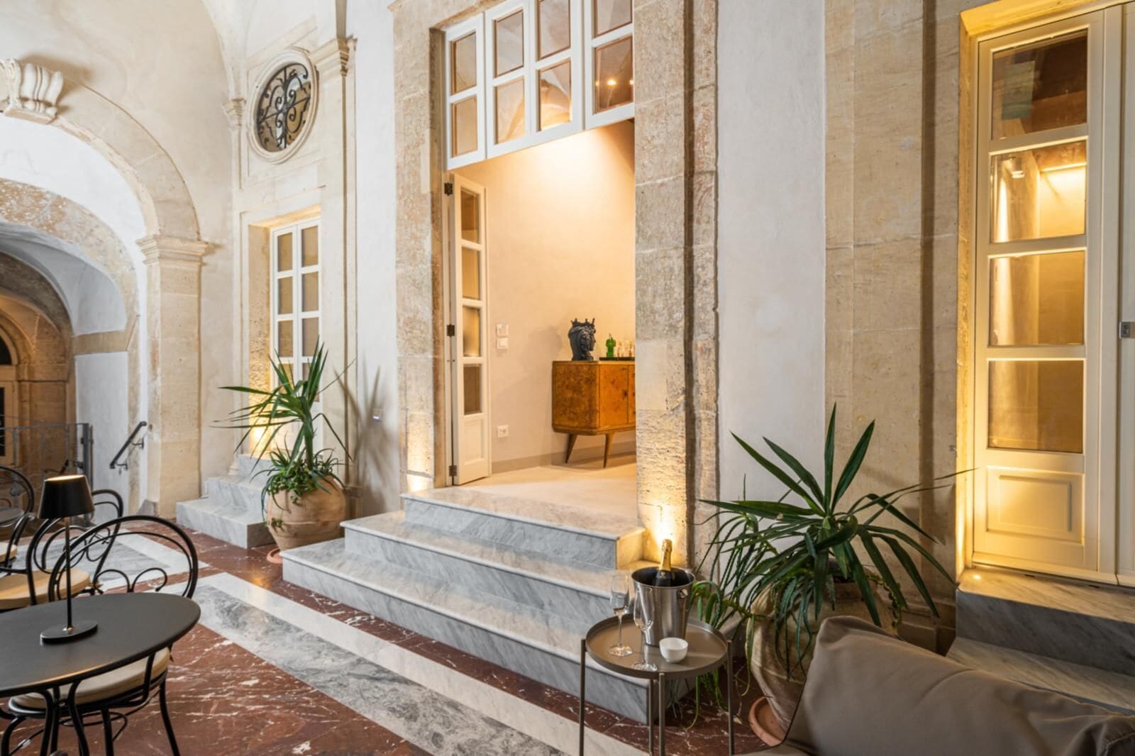 A lobby with marble flooring, a staircase leading to an upper level, a wooden cabinet, and a table with a bottle and glasses.