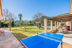 Ein Tennisplatz mit Netz, umgeben von einer überdachten Sitzgelegenheit mit Stühlen und einem Tisch, und ein Pool im Hintergrund.