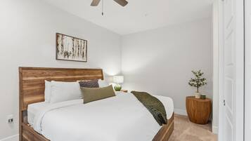 Un dormitorio con cabecera de madera, cama con ropa de cama blanca, mesita de noche con lámpara, un cuadro enmarcado en la pared y una planta en maceta.