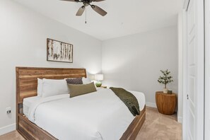 Un dormitorio con cabecera de madera, cama con ropa de cama blanca, mesita de noche con lámpara, un cuadro enmarcado en la pared y una planta en maceta.