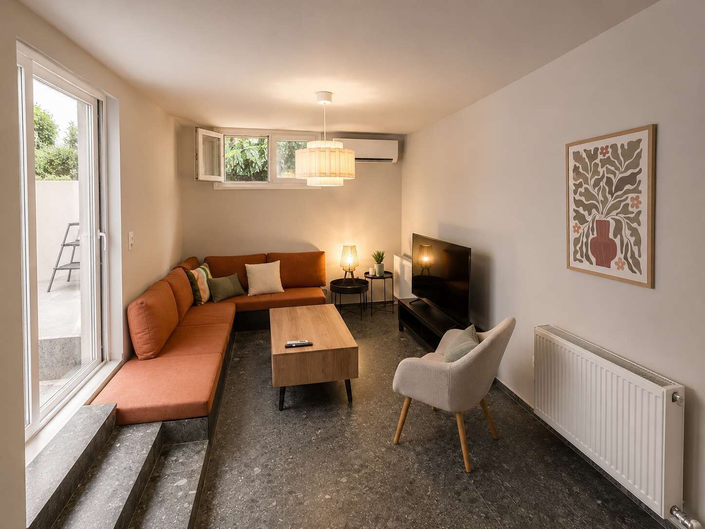 Ein Wohnzimmer mit Sofa, Couchtisch, Fernseher und Sessel. Ein Heizkörper und ein Fenster mit Ausblick sind ebenfalls vorhanden.