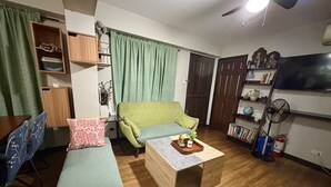 Een woonkamer met een groene bank, een houten salontafel en een televisie. Er is een boekenkast en een ventilator in de kamer.