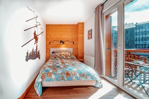 Un dormitorio con una cama, cabecera de madera, pared cubierta con plantas trepadoras, ventana con vista a edificios y balcón con mesa y sillas.