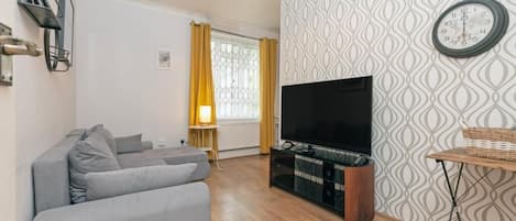 Un salon avec un canapé gris, un téléviseur à écran plat, un meuble en bois et un papier peint à motifs.