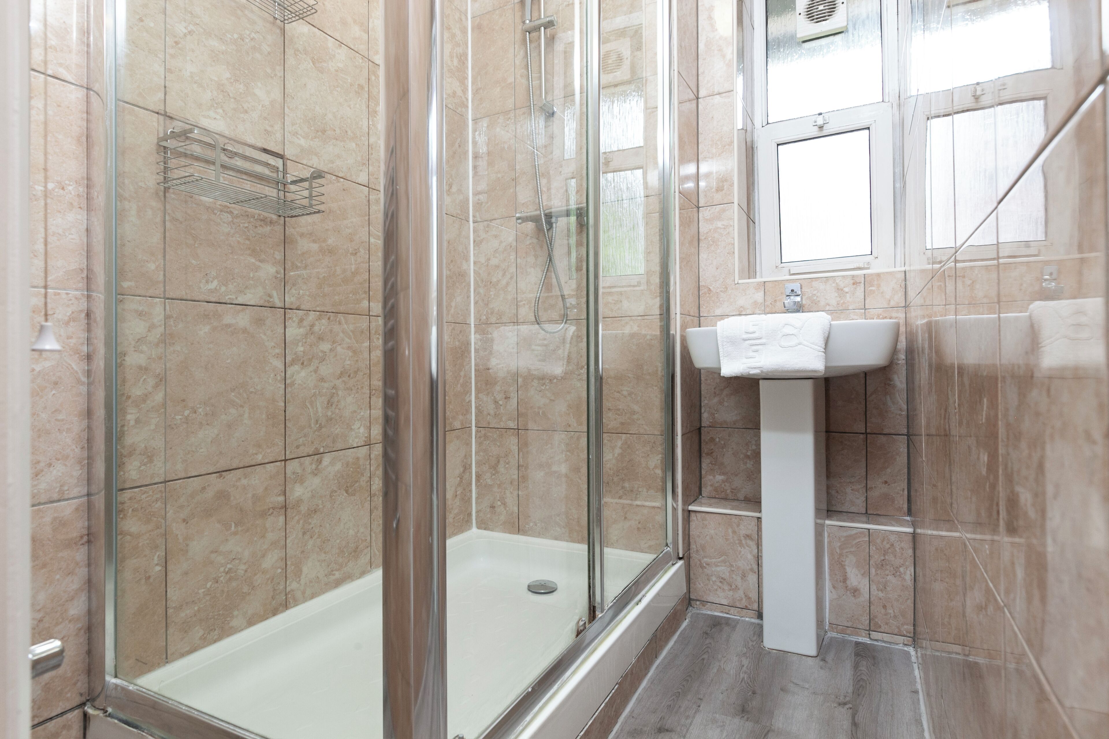 A bathroom with a glass-enclosed shower, a white pedestal sink, and beige tiled walls.