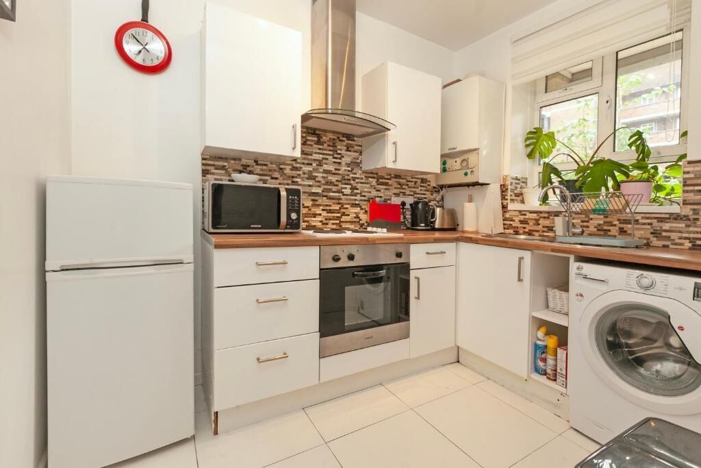 A kitchen with white cabinets, a microwave, an oven, a washing machine, and a window with plants.