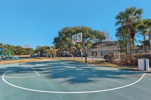 Ein Basketballplatz im Freien mit einem Korb, umgeben von Bäumen und einem Spielplatz.