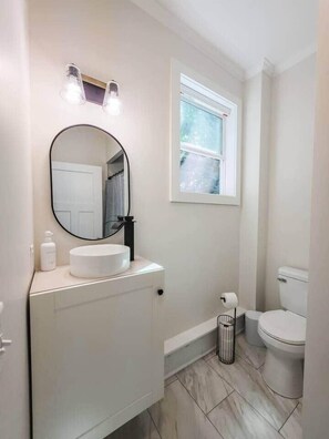 Een badkamer met een ronde spiegel, een wit wastafelmeubel, een toilet en een raam.