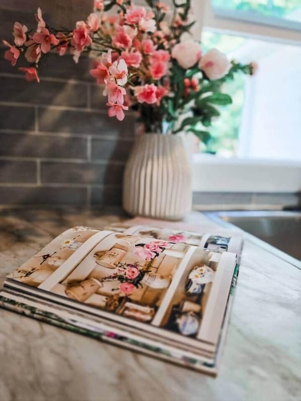 A kitchen counter with a vase of pink flowers and a stack of magazines.