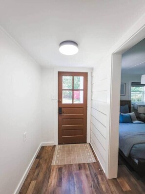 Un couloir avec une porte en bois, un paillasson à motifs et un lit avec une literie bleue dans la pièce adjacente.