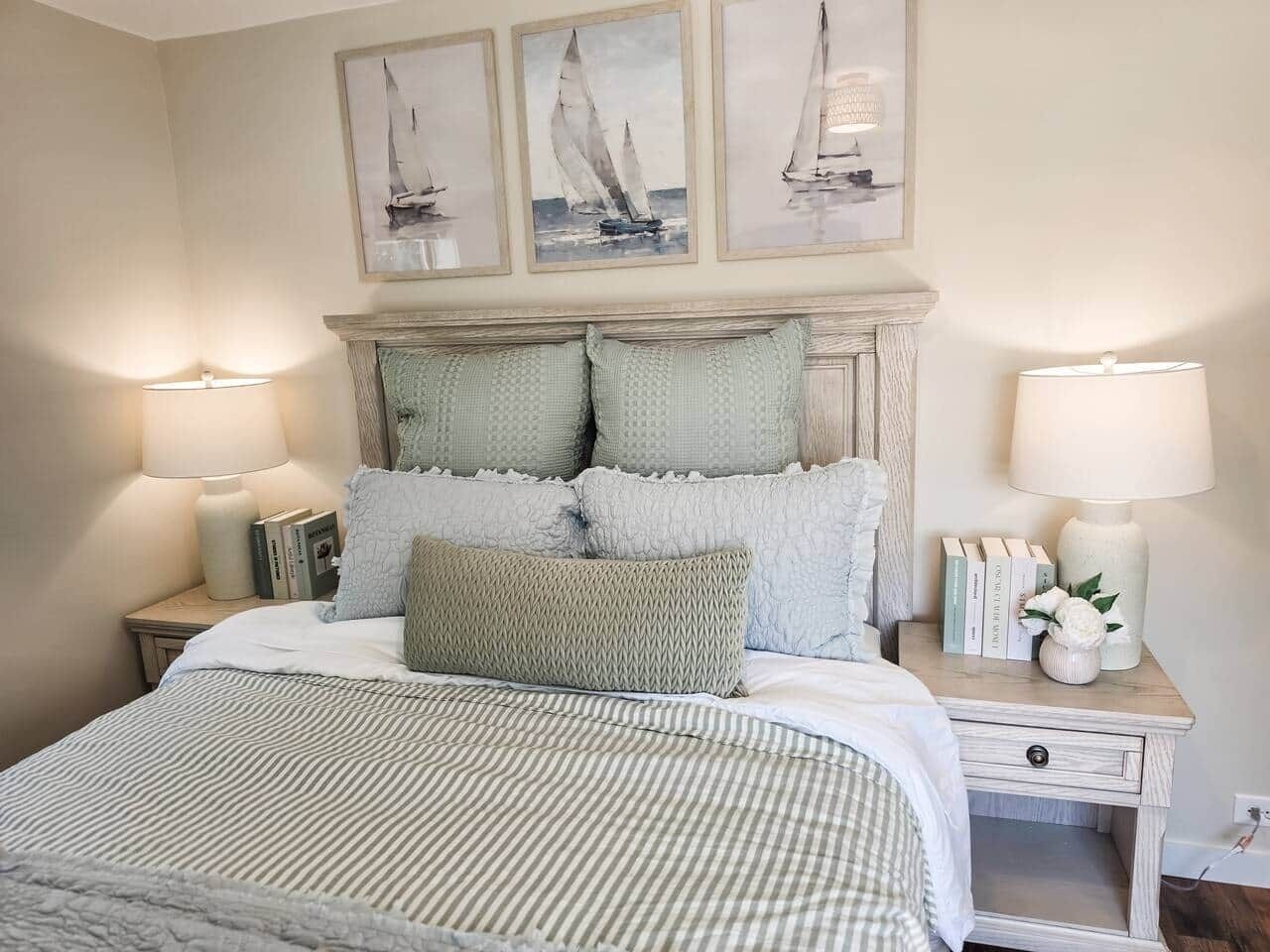 A neatly made bed with a striped bedspread, two bedside lamps, a nightstand with books and a vase, and three framed pictures above the bed.