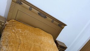 Een bed met een bedekking met gouden textuur, een houten hoofdbord met decoratieve details, en een effen muur op de achtergrond.