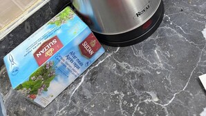Een roestvrijstalen waterkoker met zwart handvat en deksel, op een marmeren aanrecht.