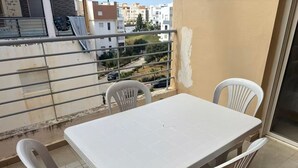 Een balkon met een witte tafel en stoelen, een metalen hekwerk en uitzicht op gebouwen en groen.