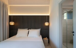 Ein modernes Hotelzimmer mit einem großen Bett, zwei Nachttischlampen, einem kleinen Nachttisch und einer Glastür zu einem Duschbereich.