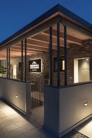 En modern byggnad med en fasad av sten, en skylt med texten ”RESIDENZA MONTE CROCIONE LAGO DI CONO” och en grind av metall.