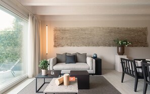 Ein modernes Wohnzimmer mit einer Couch, einem Couchtisch, einem Beistelltisch und einem Sessel. An der Wand hängt ein großes abstraktes Gemälde, und es gibt einen Blumentopf sowie eine Vase mit Blumen.