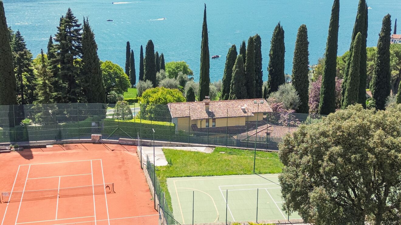 Een tennisbaan met een gebouw op de achtergrond, omgeven door bomen en met uitzicht op een waterpartij.