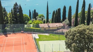 Ein Tennisplatz mit einem Gebäude im Hintergrund, umgeben von Bäumen und mit Blick auf ein Gewässer.