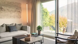A modern living room with a sofa, a coffee table, and a chair. There is a large window with a view of trees and mountains. - MC residence (Tremezzo)
