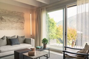 Ein modernes Wohnzimmer mit einer Couch, einem Couchtisch und einem Sessel. Ein großes Fenster bietet Blick auf Bäume und Berge.