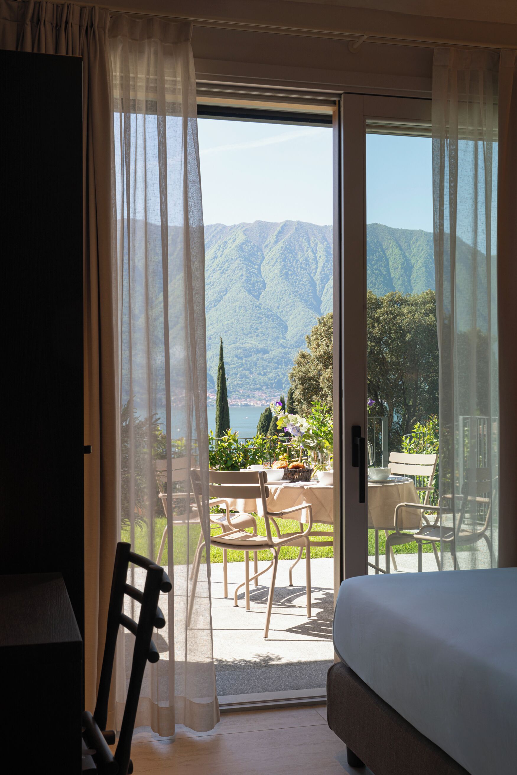 A room with a bed, a chair, and a table with a view of mountains and greenery.