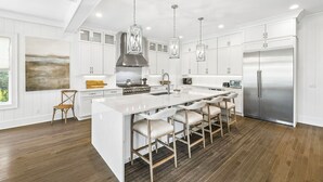 A modern kitchen with white cabinetry, a large island with bar stools, stainless steel appliances, and pendant lighting. - Elegant Pet-Friendly Coastal Retreat with Private Pool, Cabana, & Elevator (Seacrest West)