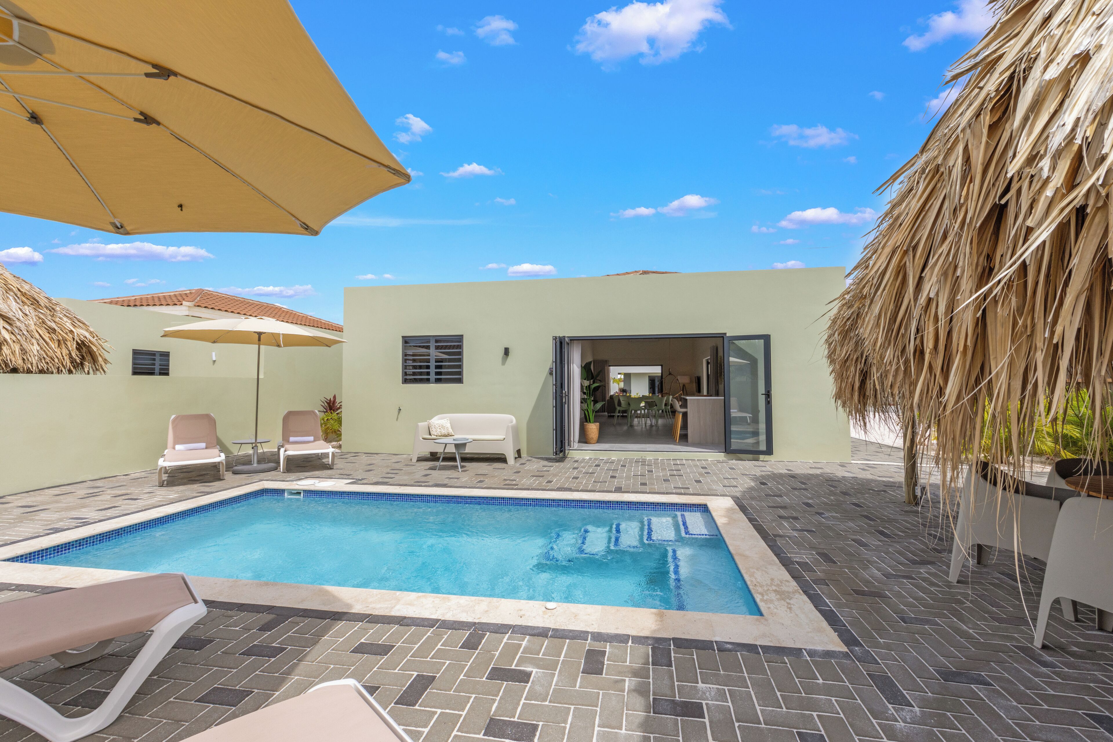 A swimming pool with lounge chairs and umbrellas, a thatched roof, and a view of the interior.